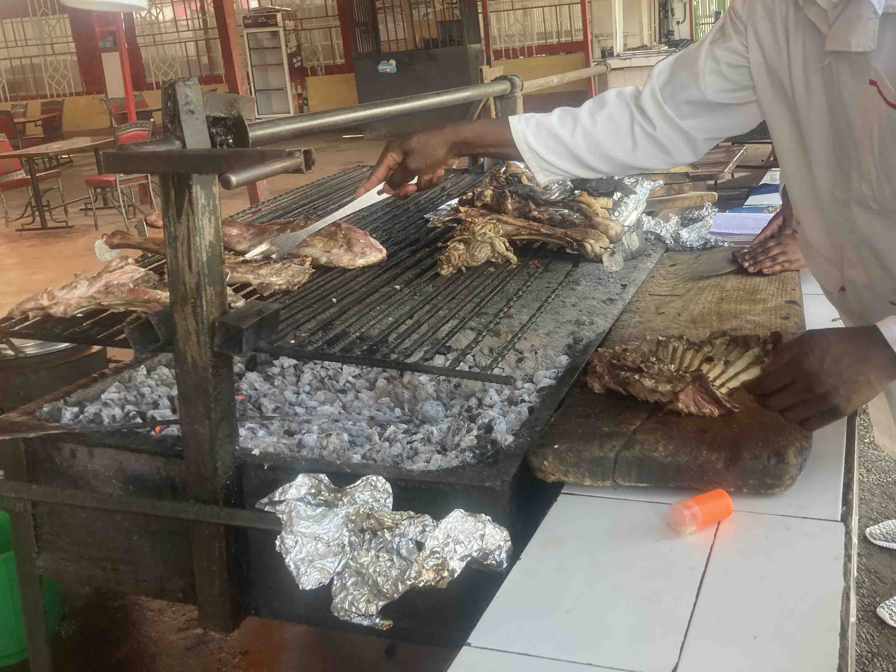 A chef grilling Nyama Choma (roasted meat) over charcoal at an open-air restaurant.