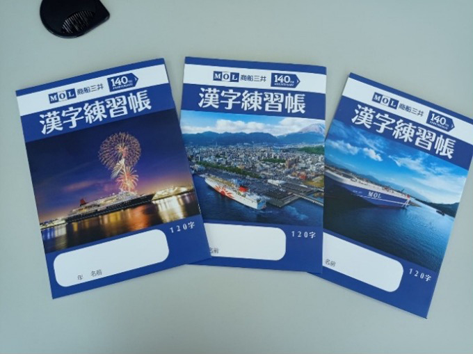 Three Kanji practice notebooks with MOL branding are displayed on a table. Each cover features different MOL ships, including a cruise ship with fireworks, a ship docked in a port city, and a cargo vessel at sea.