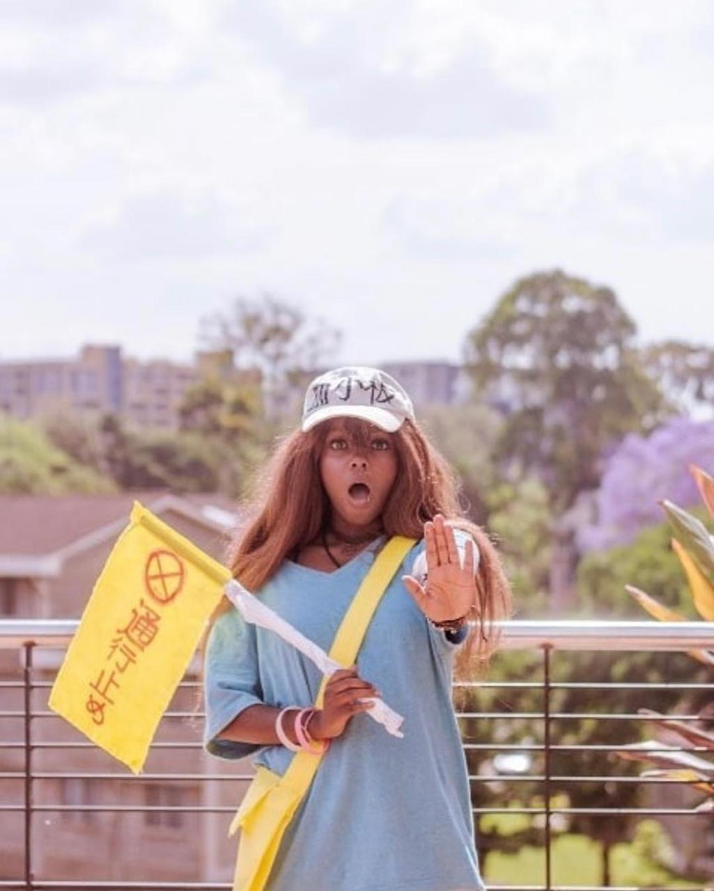 A cosplayer dressed as a traffic guide character, holding a yellow flag with Japanese text, making a stopping gesture.