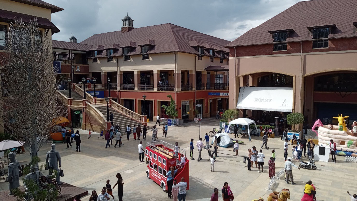 Outdoor plaza at The Hub Karen shopping mall in Nairobi, with people, shops, and a red London-style bus.