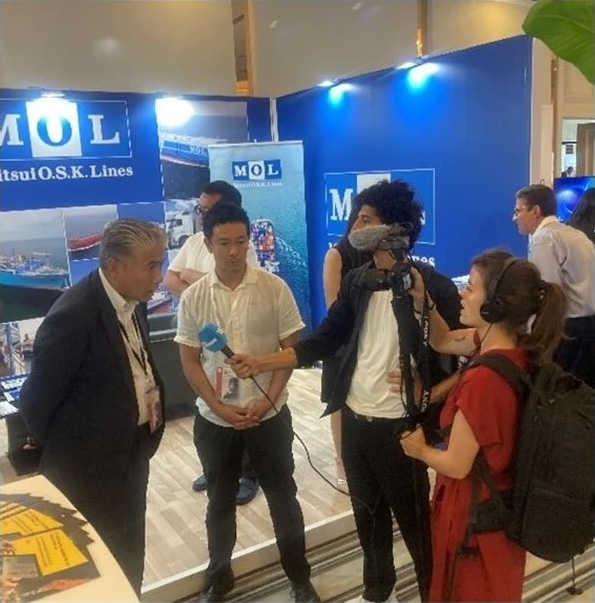 MOL staff being interviewed by a media crew at a trade show booth, with Mitsui O.S.K. Lines branding and display materials in the background.