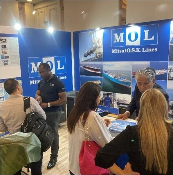 MOL representatives engaging with visitors at a trade fair booth, explaining services and displaying shipping-related visuals.