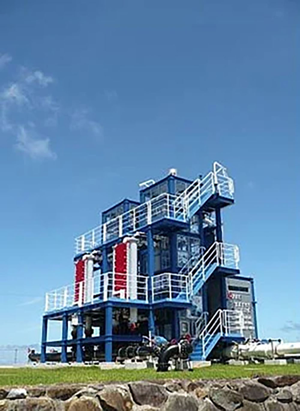 A compact, multi-level offshore or industrial test facility structure painted in blue and white, equipped with piping, tanks, and staircases, standing outdoors under a clear blue sky.