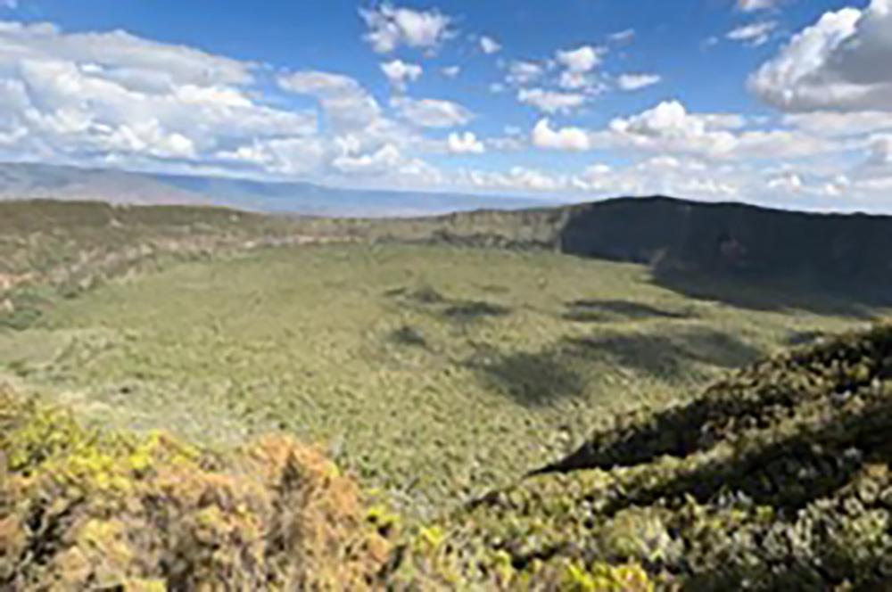 Aerial view of Mount Longonot’s massive volcanic crater in Kenya, densely covered with green forest, with blue sky and clouds above.