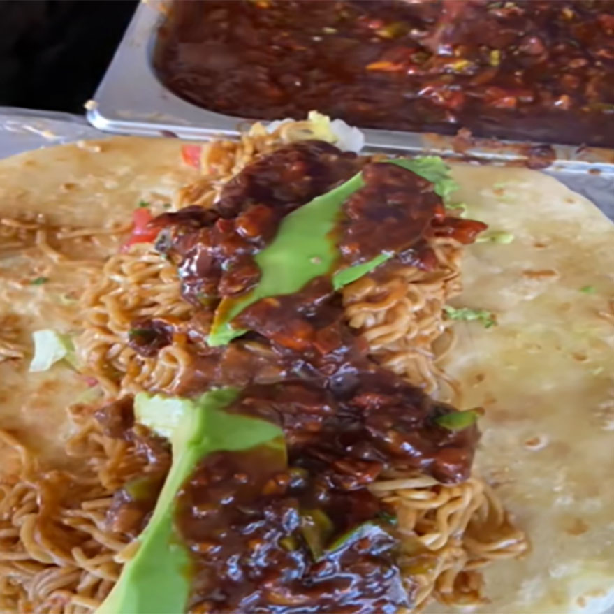 Chapati being prepared with noodles, avocado slices, and a generous topping of spicy sauce.