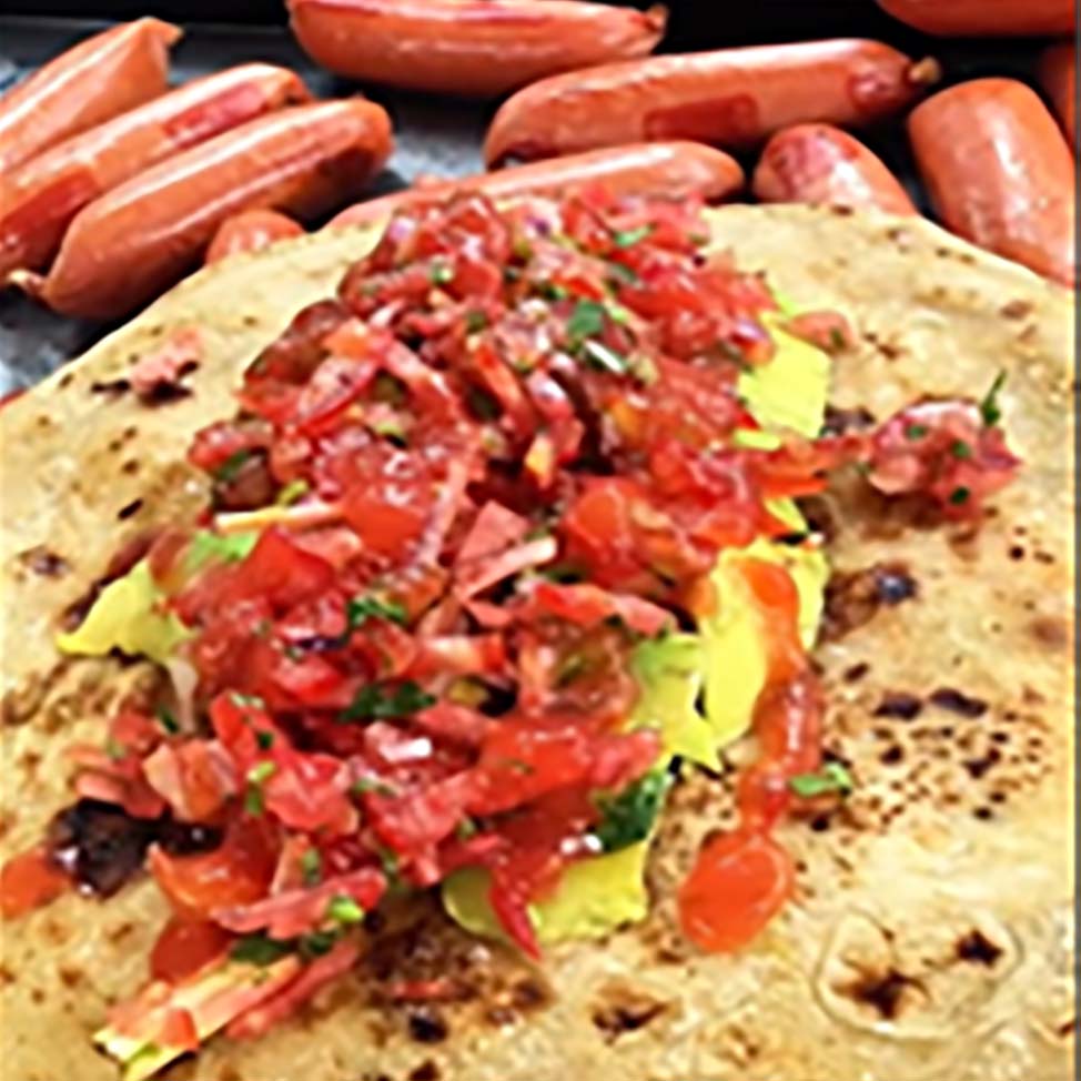 Chapati topped with avocado slices and freshly chopped kachumbari, surrounded by sausages.
