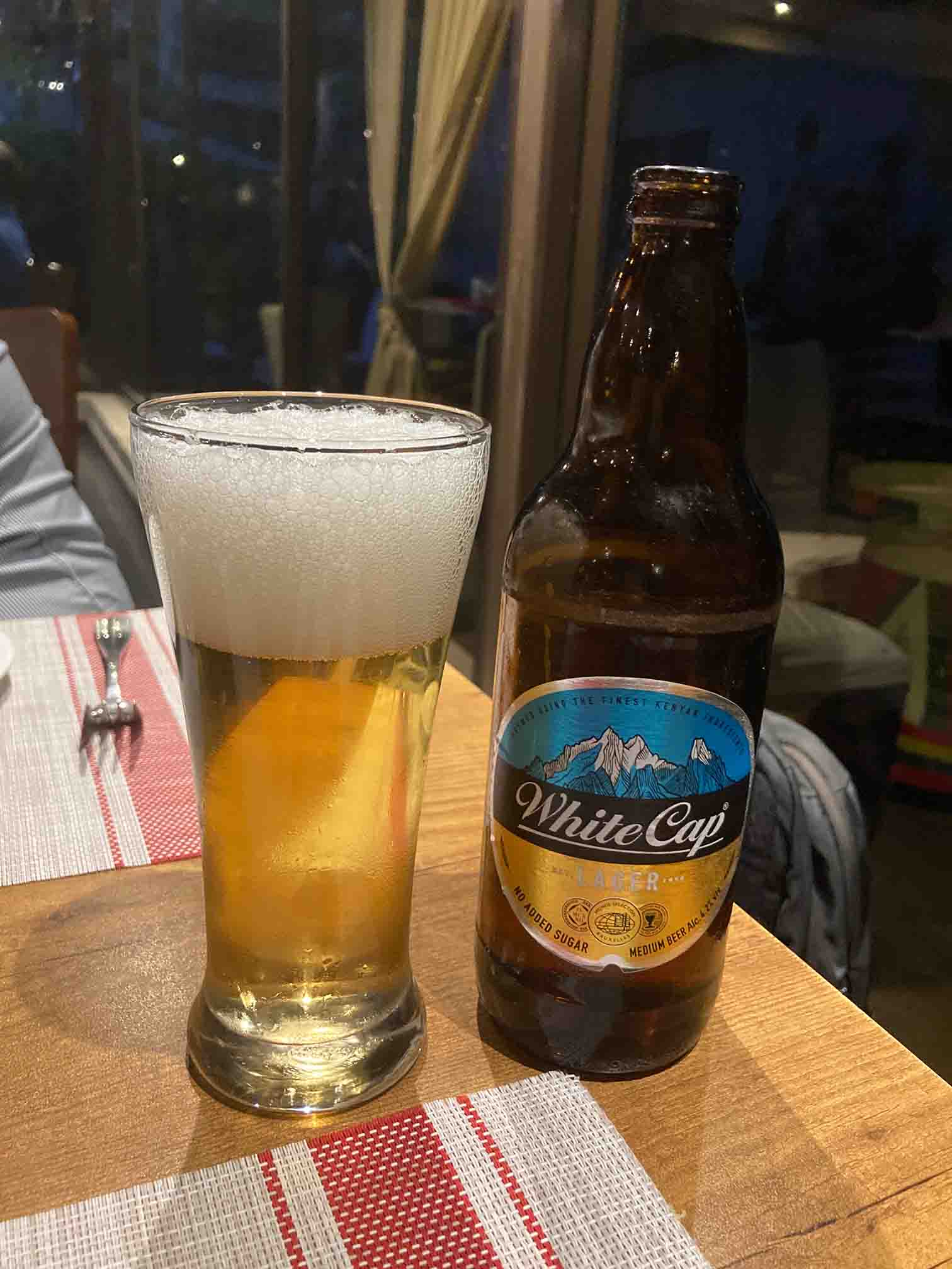 A glass of freshly poured White Cap beer with thick foam beside an opened White Cap bottle on a restaurant table.