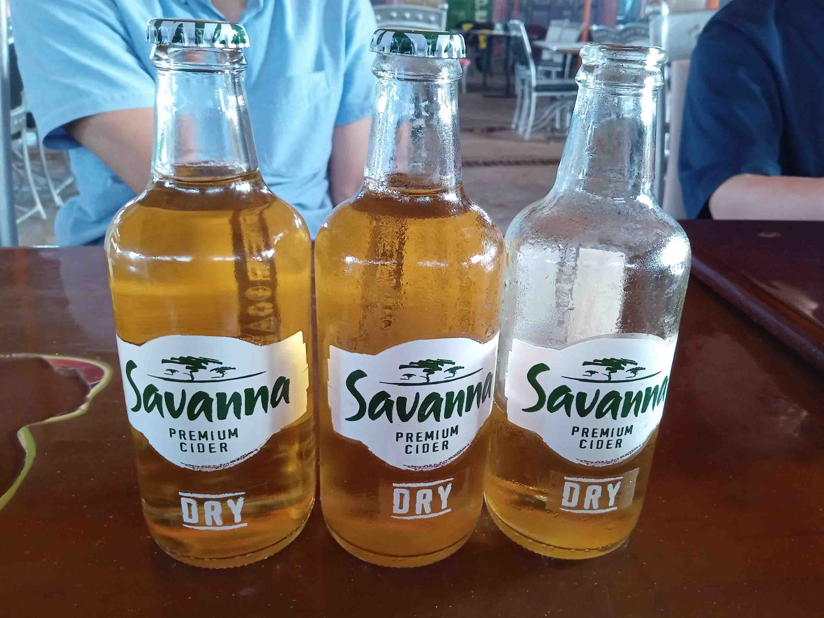 Three cold Savannah Dry cider bottles on a table, with condensation visible and people sitting in the background.