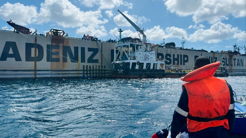 A marine operations crew member wearing an orange life jacket observes a large floating power ship on the water, with a support vessel alongside and bright clouds overhead.
