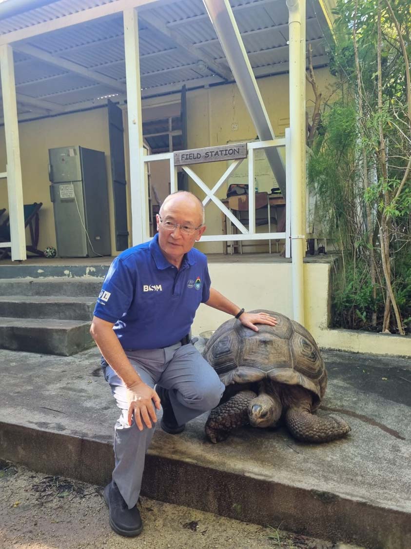 MOL CEO Takashi Hashimoto kneeling beside a large Aldabra tortoise at a conservation facility in Mauritius.