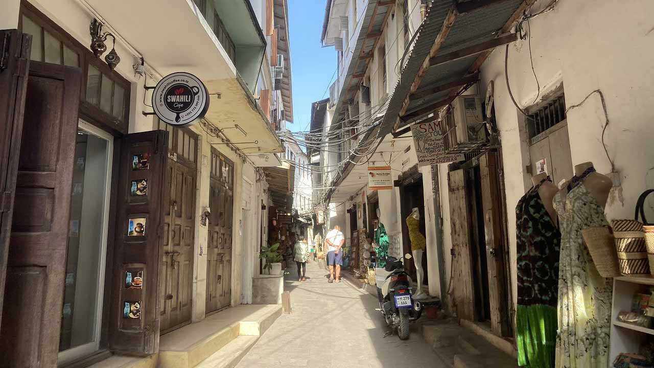 Narrow alley in Stone Town with traditional buildings, overhead cables, and small local shops.