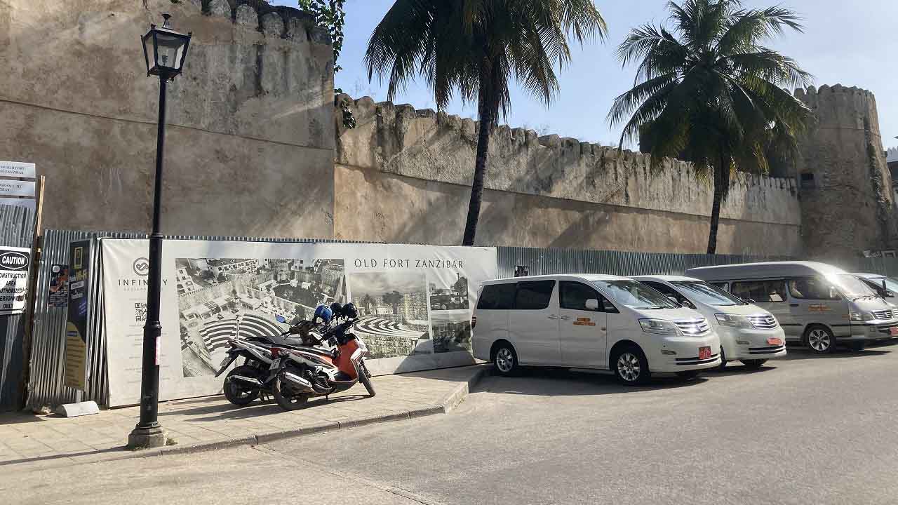 Exterior view of the Old Fort area in Stone Town with stone walls, parked vehicles, and palm trees.