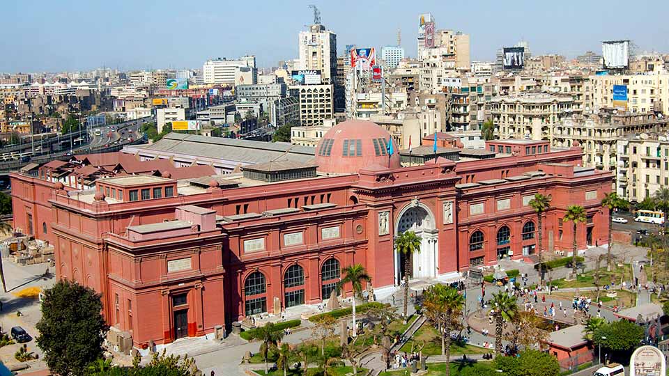 The historic Egyptian Museum in Cairo, a red neoclassical building opened in 1902, surrounded by the cityscape of downtown Cairo.