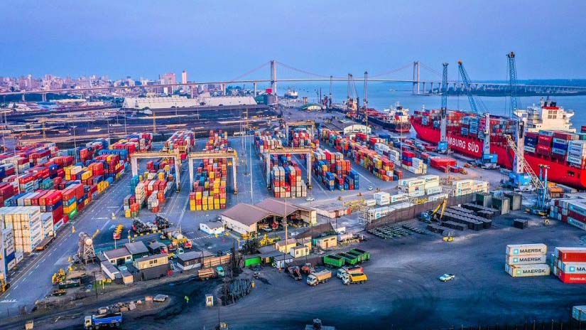 Aerial view of the Port of Maputo container terminal with stacked colorful containers, cargo cranes, trucks, and vessels operating near the waterfront, with the city and a bridge in the background.