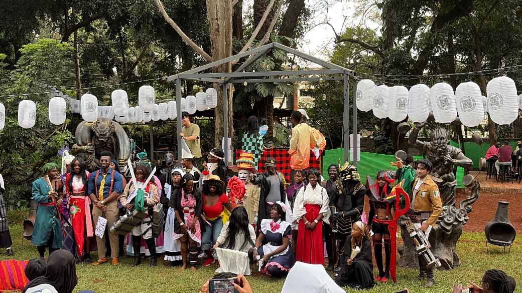 Group of cosplayers posing outdoors at a Japanese cultural festival in Kenya, with paper lanterns, statues, and a stage in the background.
