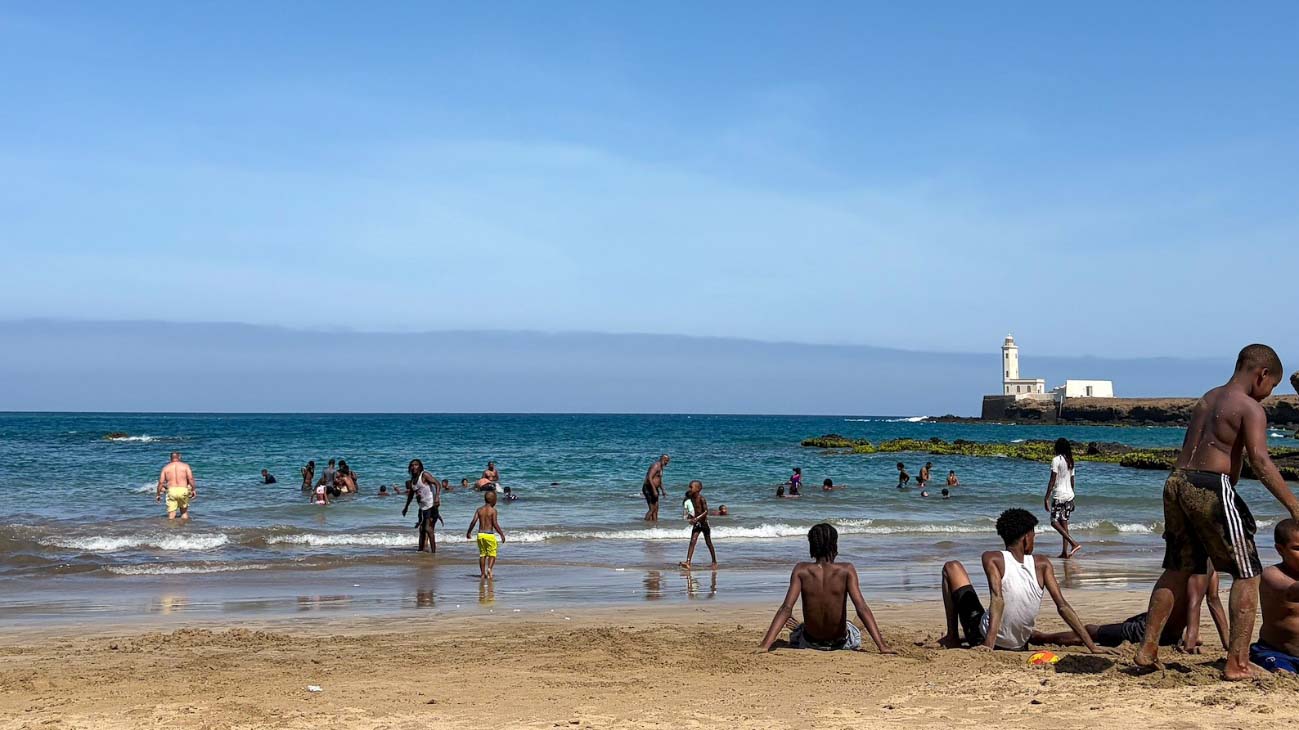 People enjoying a sunny beach in Cabo Verde, with the Atlantic Ocean and a lighthouse visible in the background.
