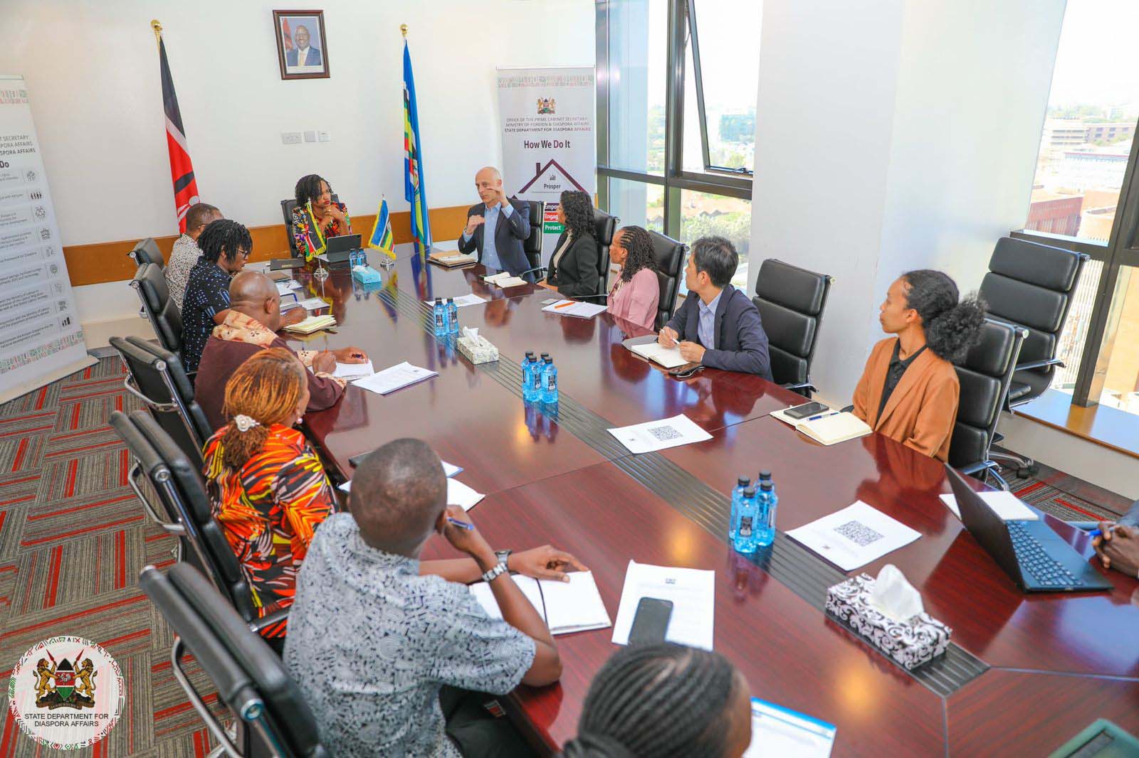 A group of professionals sits around a large conference table in a modern office, actively engaging in a discussion. Notebooks, documents, and water bottles are placed on the table. A man in a suit is speaking, while others are attentively listening. The Kenyan and East African Community flags stand in the background, along with a banner for the 'State Department for Diaspora Affairs.