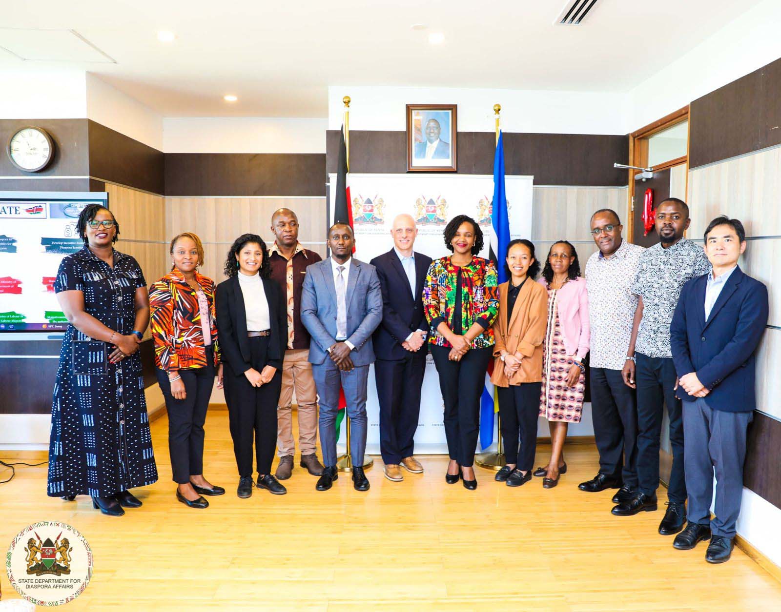 A group of twelve people, including men and women, stand together for a group photo in an office setting. They are dressed in formal and semi-formal attire, with some wearing vibrant African prints. Behind them, the Kenyan and East African Community flags are displayed, along with a framed portrait of a leader. A banner for the 'State Department for Diaspora Affairs' is visible on the left side.