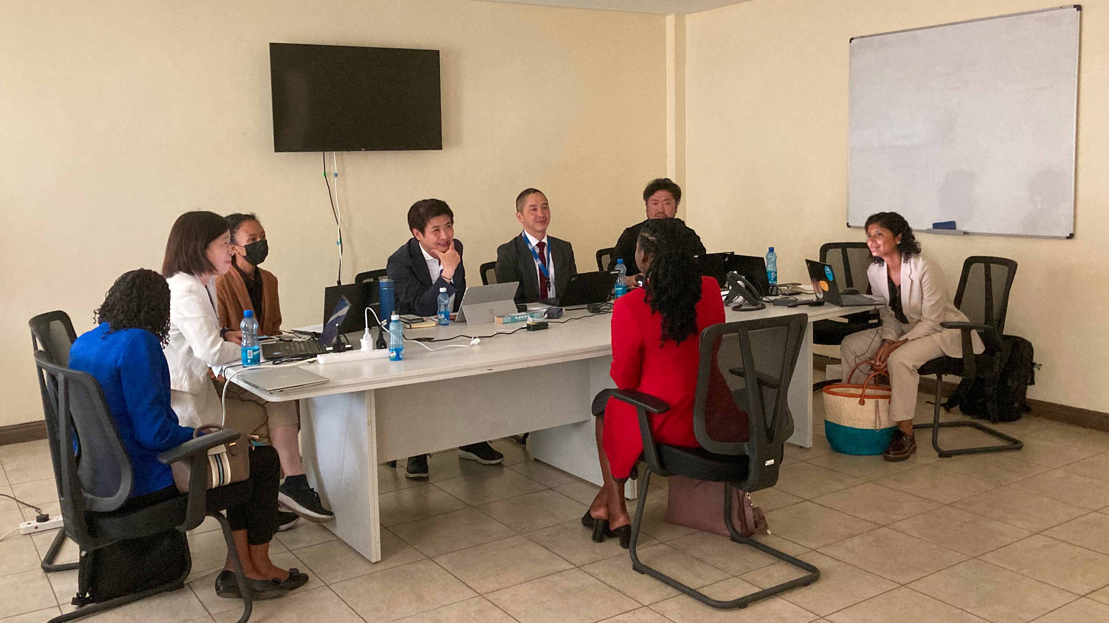 A group of professionals sits around a large conference table in a modern office, actively engaging in a discussion. Notebooks, documents, and water bottles are placed on the table. A man in a suit is speaking, while others are attentively listening. The Kenyan and East African Community flags stand in the background, along with a banner for the 'State Department for Diaspora Affairs.