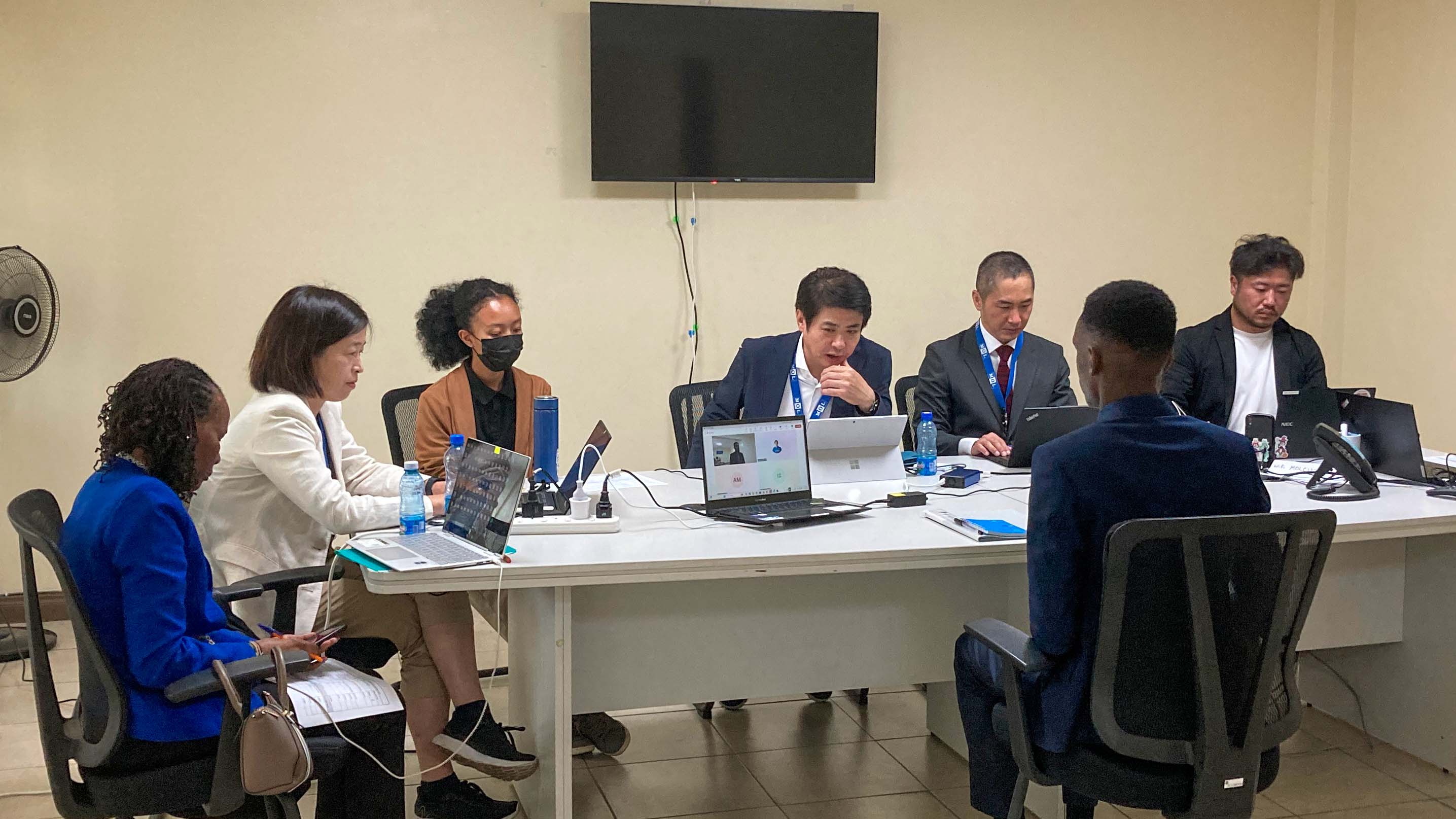 A formal interview setting with several interviewers sitting behind laptops in a meeting room. They are interviewing a male candidate dressed in a dark blue suit. The interviewers appear to be taking notes and reviewing information on their screens.