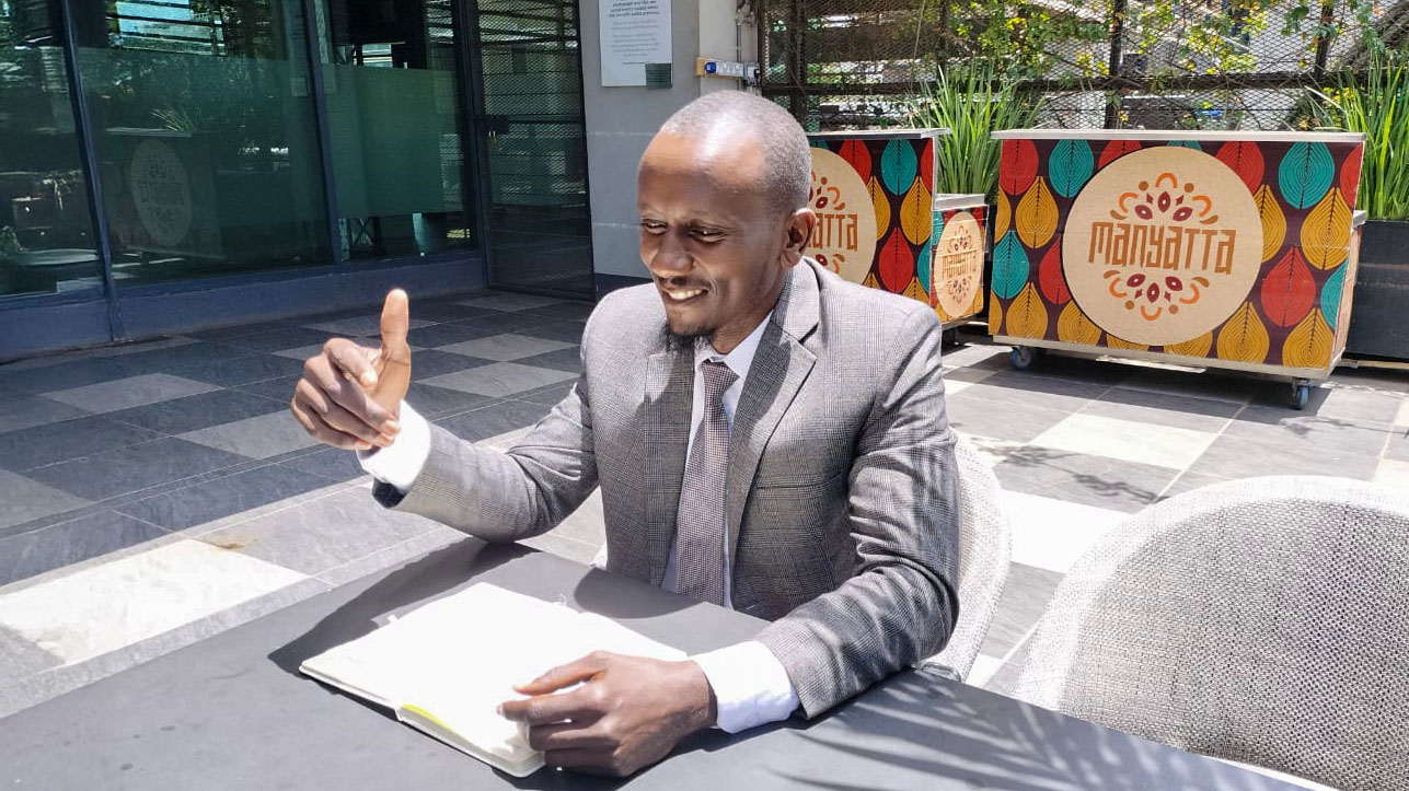 A man in a gray suit giving a thumbs up while sitting at a table outside, smiling with a notebook open in front of him.