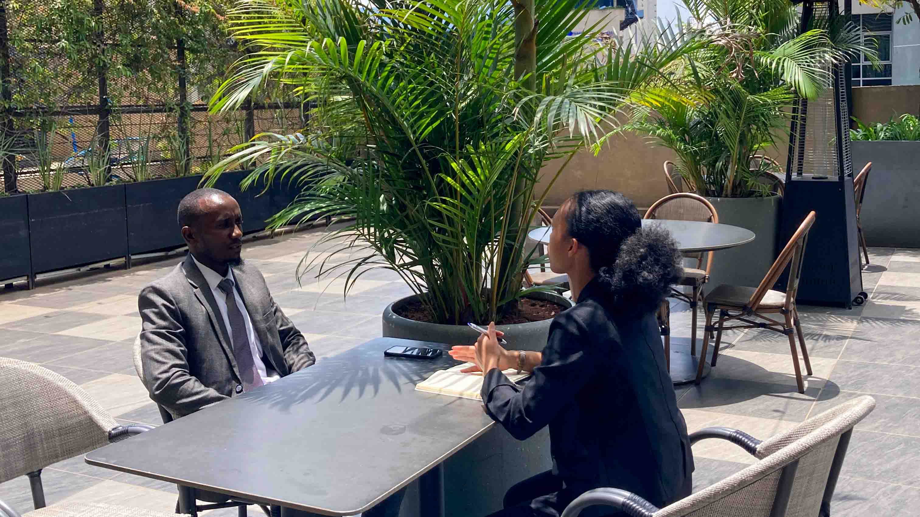 A man in a suit and a woman in black business attire having an interview at an outdoor table with large potted plants around them.