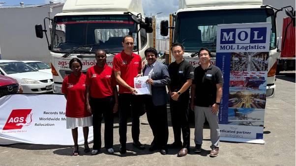A man in a suit and a woman in black business attire having an interview at an outdoor table with large potted plants around them.