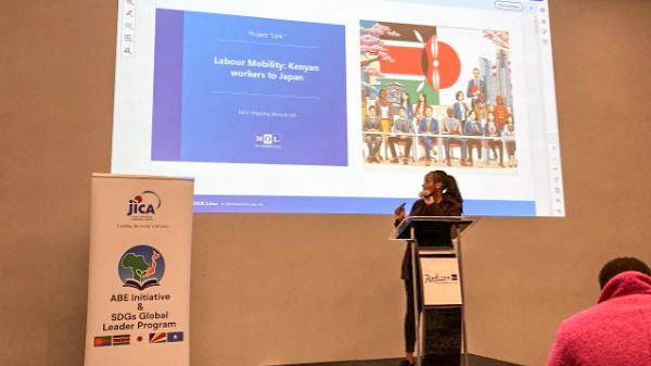 A presenter speaking at a podium in front of a screen showing a slide titled “Labour Mobility: Kenyan workers to Japan.”