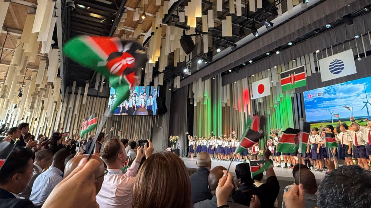 Audience waving Kenyan flags during Kenya Day at EXPO2025 in Osaka, with Japanese and Kenyan flags displayed on stage and children performing in the background.”