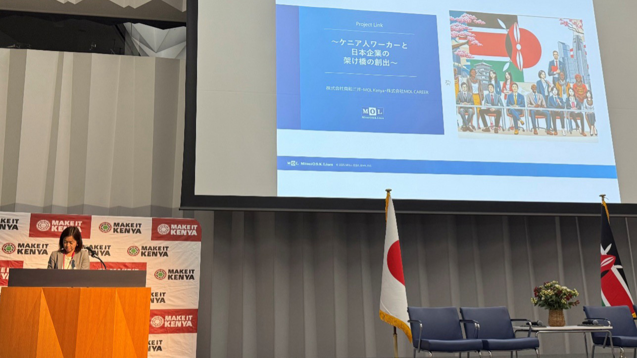 Speaker presenting on stage during a Kenya Day session at EXPO2025, with Japanese and Kenyan flags on both sides and an illustration of collaboration shown on the screen.