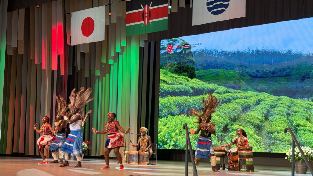 Traditional Kenyan dancers performing on stage at Kenya Day in EXPO2025, with a scenic image of green fields displayed on the screen behind them.