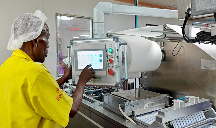 A factory worker in a yellow coat operating a touchscreen control panel on a paper packaging or medical production line, wearing a hair net for hygiene.