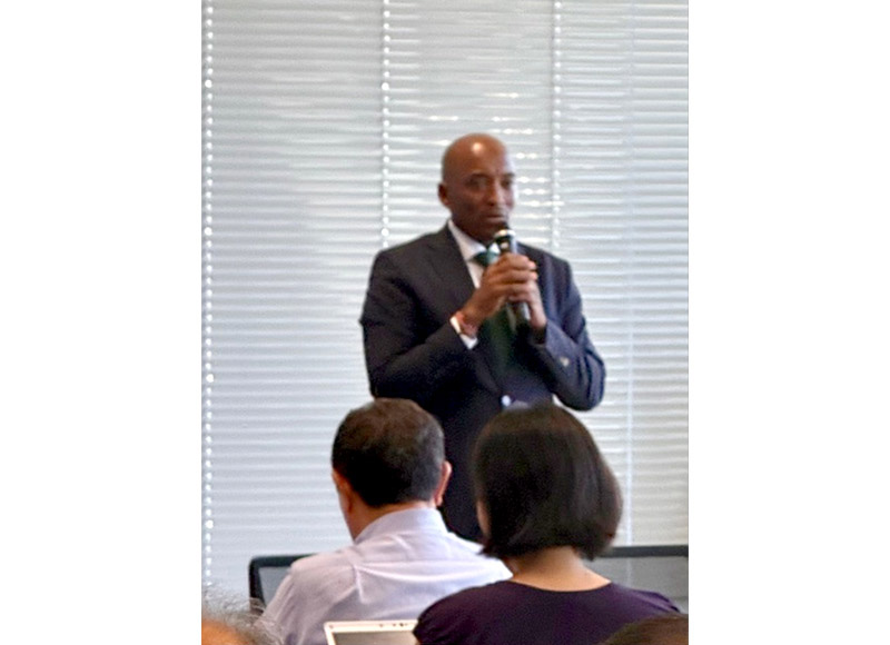 A man in a dark suit is speaking into a microphone while standing in front of an audience at a seminar.
